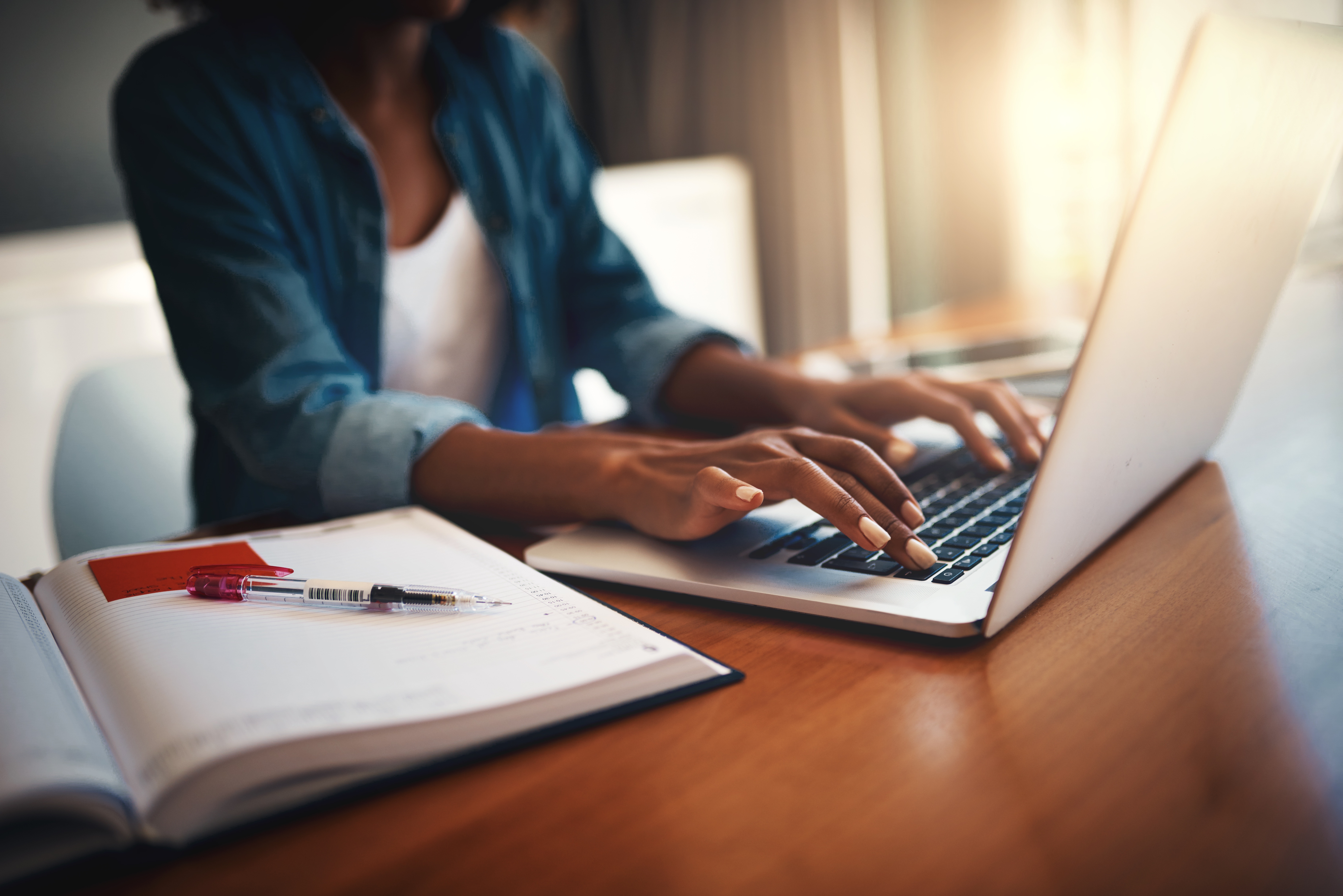 A woman sitting at a table typing on a laptop with a notebook nearby.