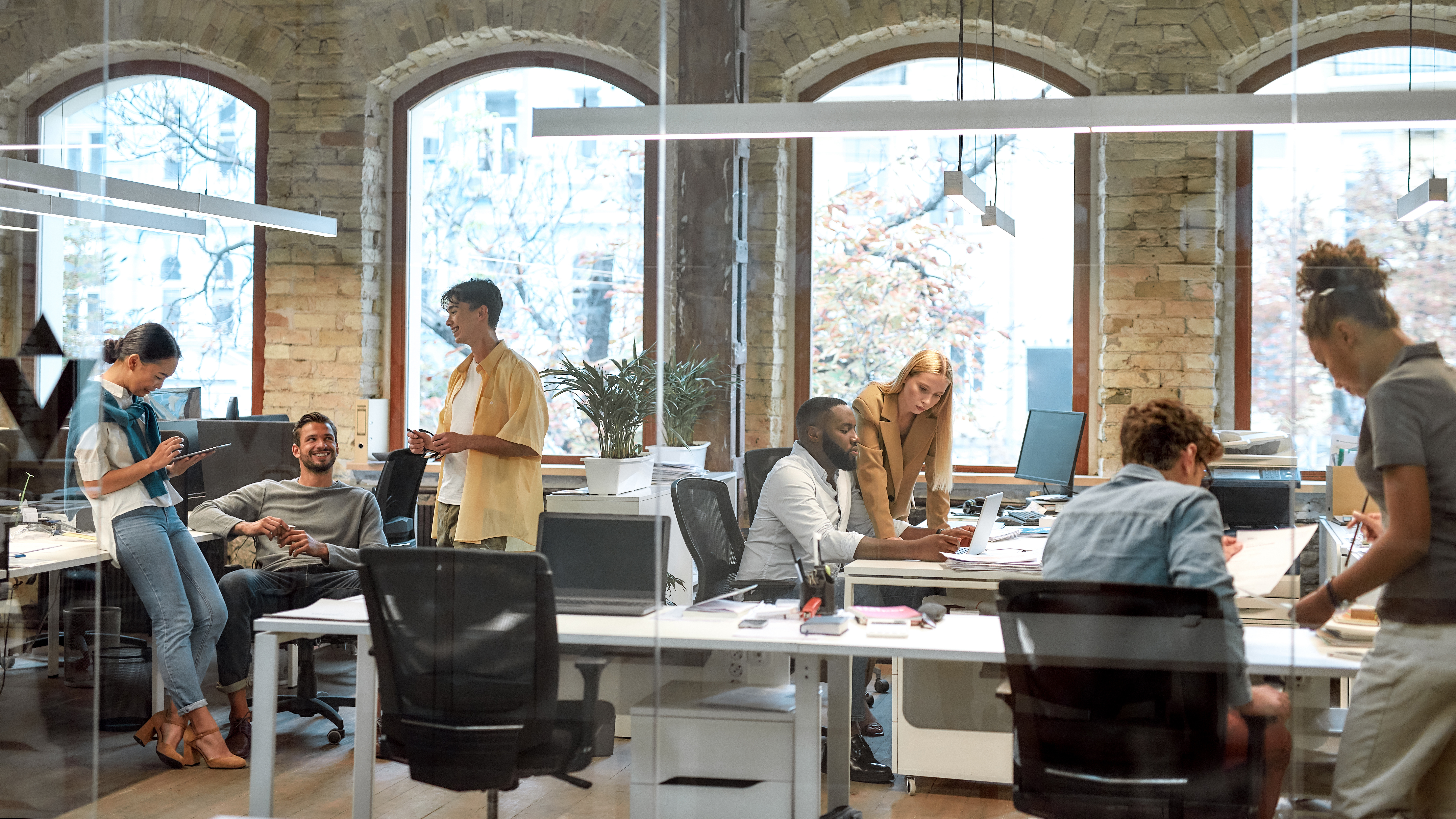 Modern open office with diverse coworkers collaborating, using computers, and discussing projects in a casual, creative workspace.